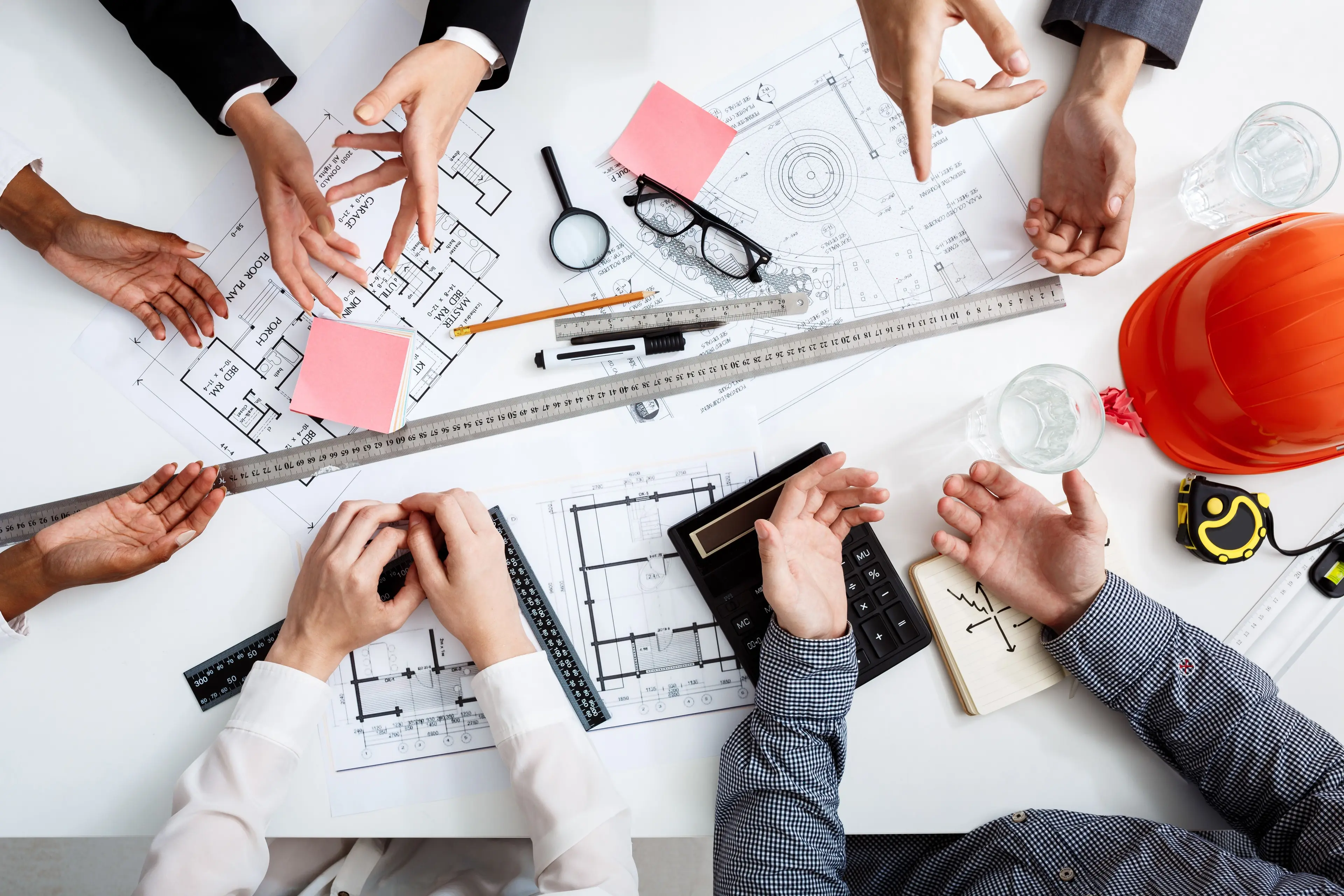 Multiple hands point at architectural blueprints and design tools on a white table.