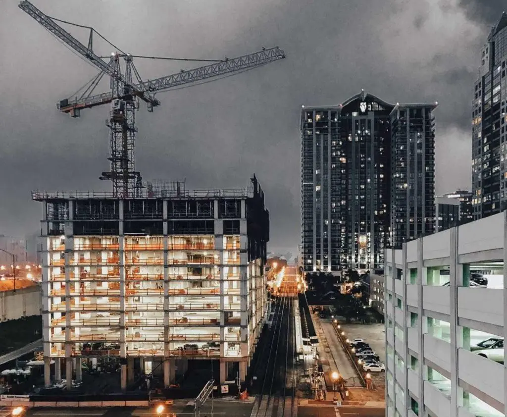 Large construction crane towers over a developing city with illuminated buildings at night.