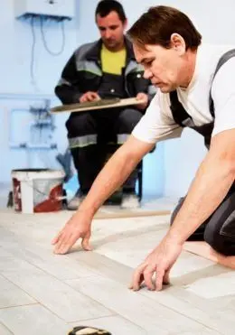 Two workers install flooring, one kneels to measure, another observes with a clipboard.