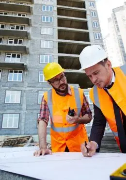 Two construction workers in hard hats review blueprints at a building site.